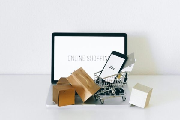 حاسوب محمول يظهر عليه نص "Online Shopping" وبجواره عربة تسوق صغيرة تحتوي على هاتف يعرض كلمة "Pay" وبطاقات دفع، مع أكياس وصناديق تسوق، في مشهد يرمز للتسوق والدفع عبر الإنترنت.